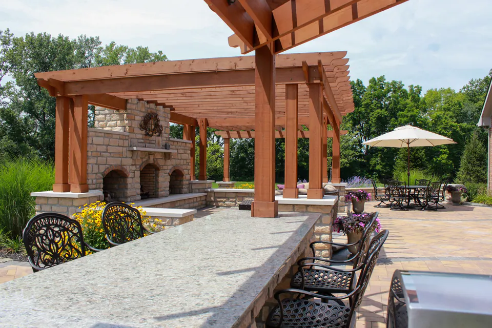 Stone bar counter looking back toward wood pergola, outdoor fireplace, paver patio, dining table, and planter pots.
